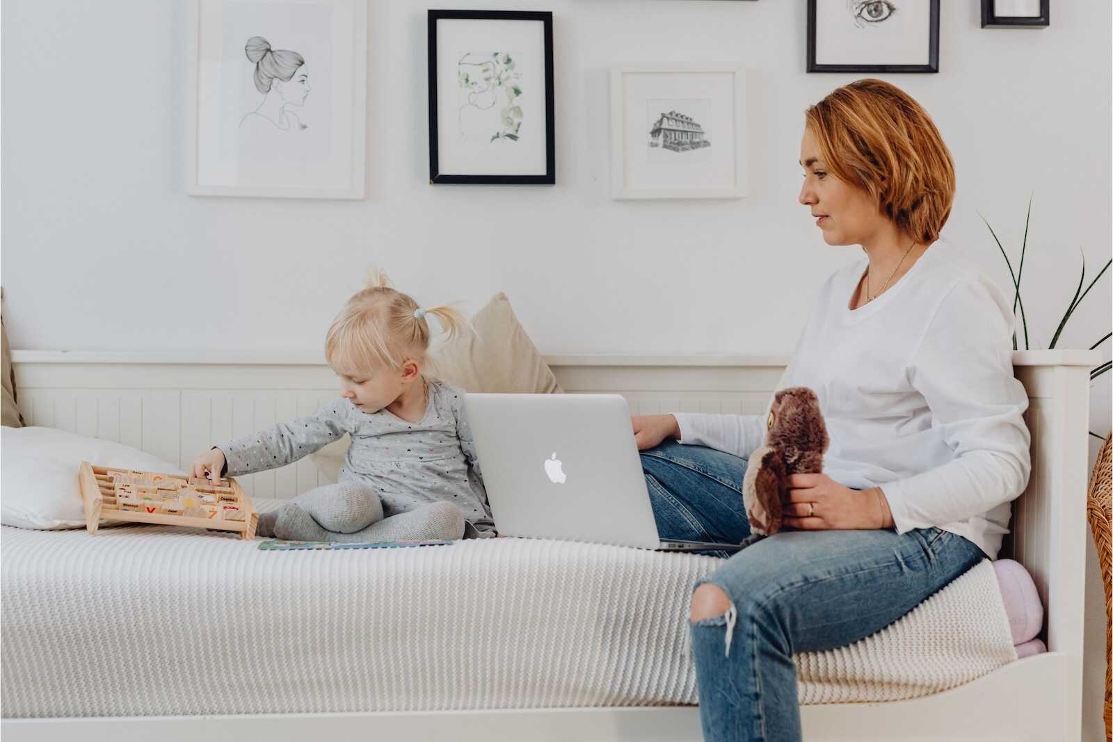 Mom sitting on a bed with her child while working on a laptop, illustrating daily routines for moms balancing work and parenting.