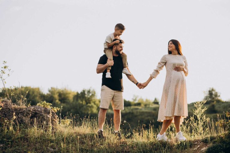 Expectant parents walking outdoors with son on dad’s shoulders during family maternity photos.