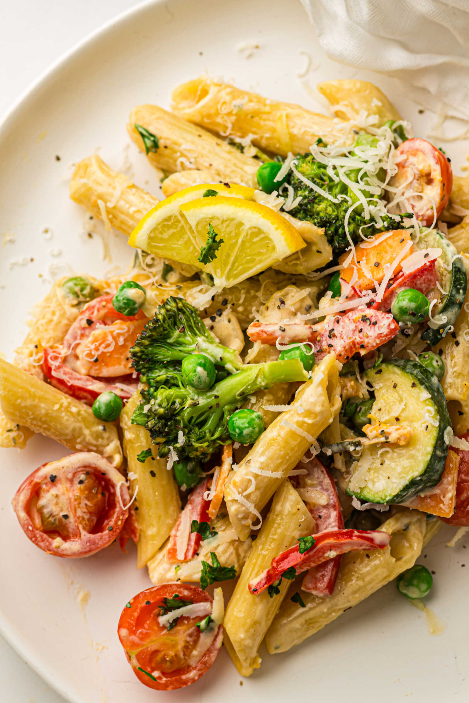 Creamy pasta primavera with penne, broccoli, zucchini, peas, cherry tomatoes, and lemon garnish on a white plate.