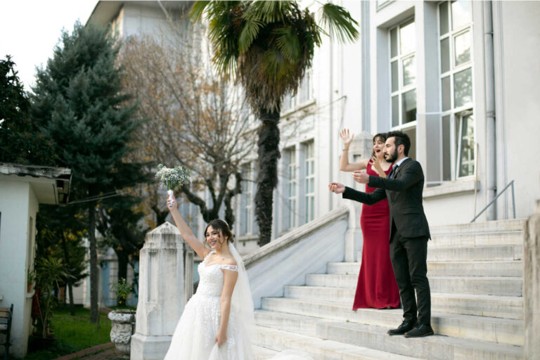 Bride celebrates outside with her bouquet while wedding guests cheer on the steps of a grand venue.